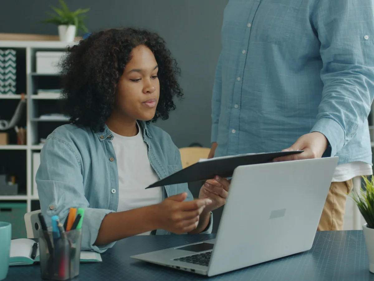 Billing specialist at a laptop reviewing claim paperwork handed over by a colleague